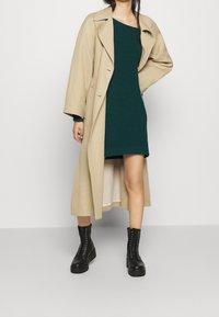 Trench coat beige sopra un vestito a coste verde scuro, abbinato a stivaletti neri con lacci alla caviglia. Il cappotto presenta una vestibilità ampia e maniche larghe.