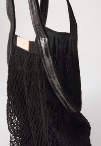 Borsa tote in rete nera con texture intrecciata, interno nero liscio e etichetta in pelle con testo in rilievo. Le maniglie sono piatte e resistenti.