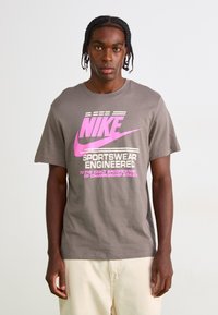 Pilkas medvilnės marškinėliai su rožiniu Nike logotipu ir tekstu su horizontaliomis juostelėmis. Trumpomis rankovėmis ir laisvu kirpimu. Nėra matomų raštų.