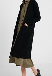 Lång svart cardigan med öppen framsida, ribbade manschetter och fickor, lager-på-lager över en grön skjortklänning med delad nederkant. Slät textur.