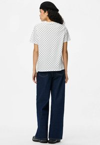 Camiseta blanca de manga corta con lunares negros, combinada con unos pantalones vaqueros oscuros anchos. Boina negra de perfil bajo llevada en la cabeza.