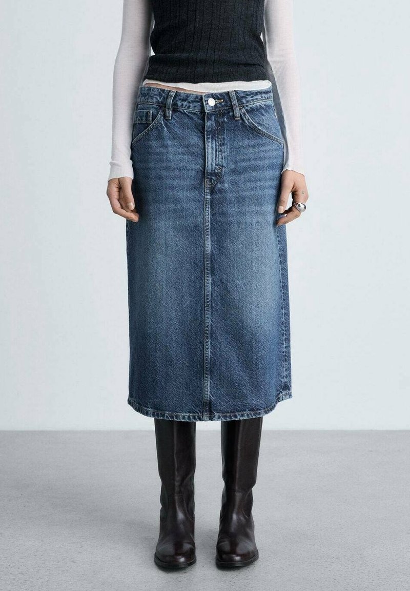 Femme portant une jupe midi en denim bleu, des bottes hautes noires, un haut noir sans manches par-dessus une chemise blanche à manches longues, debout sur un sol gris.