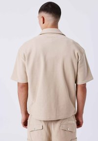 Camicia beige a maniche corte con colletto, realizzata in tessuto testurizzato. Presenta un piccolo logo sulla parte superiore della schiena. Abbinata a pantaloni beige coordinati.