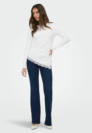 Nainen, joka pukeutunut valkoiseen pitsikoristeiseen pitkähihaiseen toppiin, tummansinisiin suoraan lahkeisiin farkkuihin ja valkoisiin teräväkärkisiin korkokenkiin, poseeraa yhdellä kädellä lantiolla.