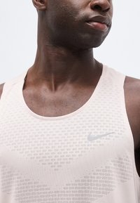 Homme noir portant un haut de sport sans manches Nike rose clair avec de petits motifs perforés sur fond blanc.