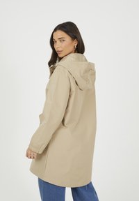 Beżowy płaszcz przeciwdeszczowy z kapturem, z elastycznymi mankietami, luźny krój i przednie kieszenie, w połączeniu z niebieskimi dżinsami. Gładka tekstura materiału.