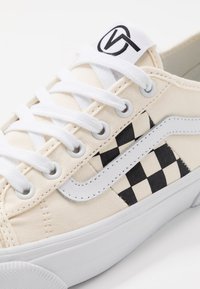 Beige canvas sneaker with white laces and a black checkerboard pattern on the side; features a white leather accent and rubber sole.