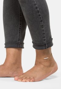 Gull armbånd med små hvite perler, kombinert med oppbrettede mørkegrå denimjeans og barføtt. Fokuser på ankel detaljen.