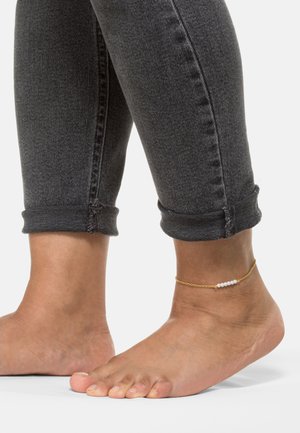 Goldenes Fußkettchen mit kleinen weißen Perlen, kombiniert mit hochgekrempelten dunkelgrauen Jeans und barfuß. Fokus auf das Detail des Knöchels.