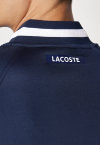 Pólo azul-marinho com um design ajustado, apresentando um colarinho riscado a branco. Etiqueta com "LACOSTE" na parte de trás está em destaque.