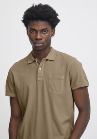 Camisa polo color caqui con mangas cortas, cuello y un bolsillo en el pecho. Tejido texturizado; presenta un detalle de doble costura en el cuello y las mangas.