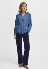 Blue patterned blouse with a V-neck, long sleeves, and loose fit, paired with navy corduroy pants and brown heeled boots.