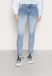 Jeans ajustados de color azul claro con una rodilla desgastada, cinco bolsillos y cierre con cremallera. Combinados con zapatillas en blanco y negro. Textura de denim suave.
