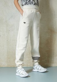 Krémové tepláky s texturovaným povrchem, černým třípruhým akcentem blízko manžety a malým černým logem Adidas na boku.