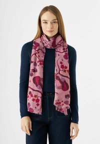 Écharpe rose ornée de motifs floraux et abstraits, fabriquée en tissu doux, avec un bord à franges, portée sur un pull noir.