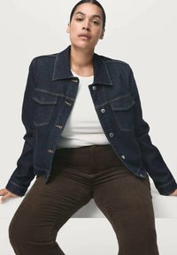 Denimjacket met een donkere wassing, voorzien van een klassieke kraag, voorzakken en knoopsluitingen. Draagt over een wit shirt en een bruine broek.