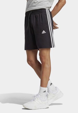 Shorts neri Adidas con strisce bianche lungo i lati e una cintura elastica con cordoncino. Il modello indossa calzature e calzini bianchi.