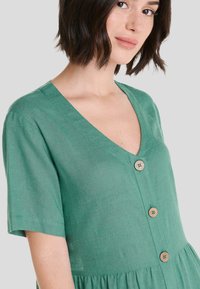 Vestido de lino verde con mangas cortas, escote en V, botones de madera en la parte delantera y cintura fruncida para añadir textura.