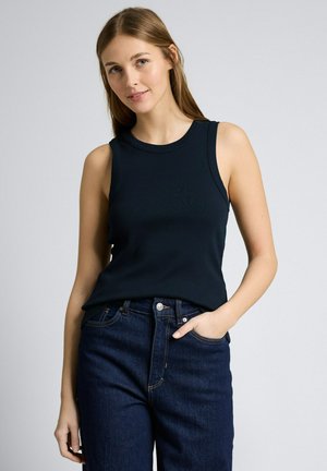 Junge Frau mit hellbraunen Haaren trägt ein schwarzes, ärmelloses Top und dunkelblaue Jeans, posiert mit einer Hand in der Tasche vor schlichtem Hintergrund.