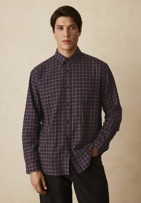 Lilla ternet skjorte med krave, der har sorte knapper og lange ærmer, lavet af et blødt, tekstureret stof.