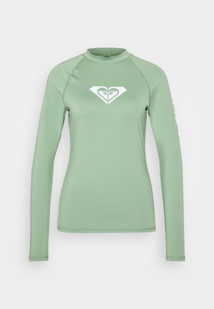 Ilgųjų rankovių rash guard šviesiai žalios spalvos audinyje. Ant krūtinės yra baltas logo, o kairėje rankovėje yra tekstinis akcentas. Sklandus, elastingas audinys.