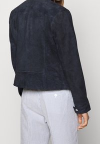 Tmavě námořnická semišová bunda s elegantním designem, viditelným šitím a jednoduchým zapínáním na cuffu. Nositá přes pruhované světlé kalhoty.