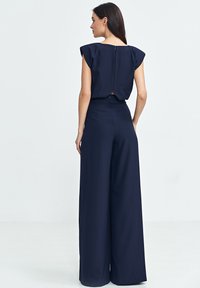 Granatowy kombinezon z szerokimi nogawkami, dopasowaną talią i rękawami typu kimono. Posiada widoczny zamek błyskawiczny na plecach oraz gładki, teksturowany materiał.