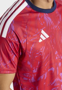 Chemise de sport rouge avec un motif dynamique violet. Présente des rayures blanches sur les épaules et un col bleu marine. Matière en polyester, manches courtes.