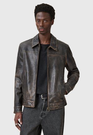 Veste en cuir marron avec une texture usée, dotée d'un col classique, d'une fermeture éclair à l'avant et de poches latérales. Associée à un T-shirt noir.