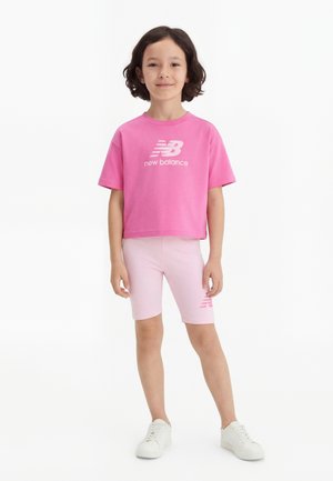 Bambino con capelli corti e scuri indossa una maglietta rosa New Balance, pantaloncini corti rosa chiaro e scarpe da ginnastica bianche su uno sfondo bianco.