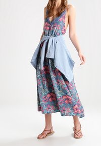 Robe maxi fleurie avec une base bleue et des fleurs roses et rouges, décolleté en V, bretelles ajustables et une chemise en denim nouée à la taille.