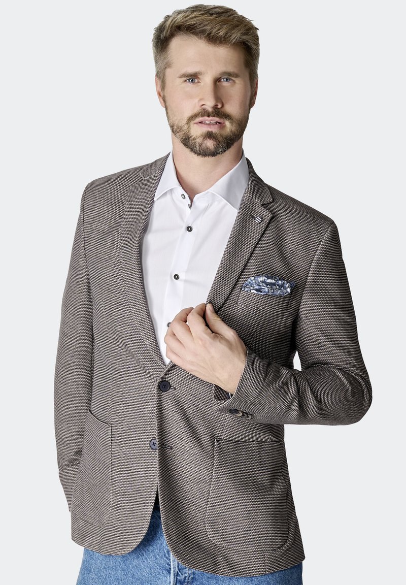 Brauner Hahnentritt-Blazer mit zwei großen Fronttaschen, blauer Futter im Brusttasche, kombiniert mit einem weißen Hemd und blauen Jeans.