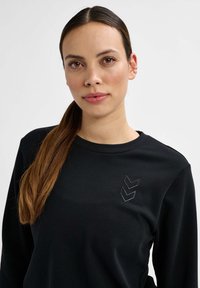 Zwarte lange mouwen top gemaakt van een katoenmix met een ronde hals. Heeft een subtiele chevron borduursel in grijs op het linkerborstgebied.