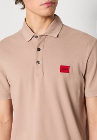 Beżowa koszulka polo z teksturowanej tkaniny, z dwoma guzikami i małą czerwoną prostokątną łatką z logo "HUGO" na lewej piersi.