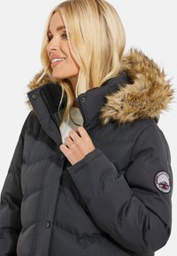 Sort pufferjakke med faux fur hætte, quiltet design og et logo patch på ærmet. Materialet virker glat og isoleret.