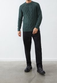 Pullover in maglia verde scuro con scollatura rotonda e polsini a coste, abbinato a pantaloni neri e scarpe nere chunky. Dettaglio del tessuto texturizzato.