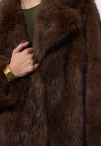 Manteau en fausse fourrure marron avec une texture moelleuse et un large col. Une main tient le tissu, mettant en valeur la riche couleur et la douceur au toucher. Bracelet en or visible.