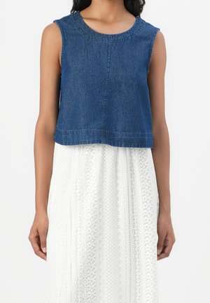 Mouwloos blauw denim cropped top, gedragen over een mouwloze witte kanten jurk, gedragen door een staand persoon met armen ontspannen langs het lichaam.