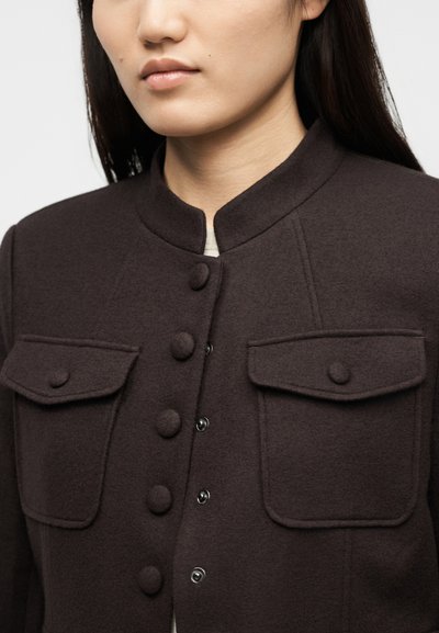 Γυναίκα που φοράει σκούρο καφέ σακάκι με κουμπιά, με τσέπες με καπάκια στο στήθος και στρογγυλή γιακά, εμφανιζόμενο από τον αυχένα έως το μέσο του κορμού.