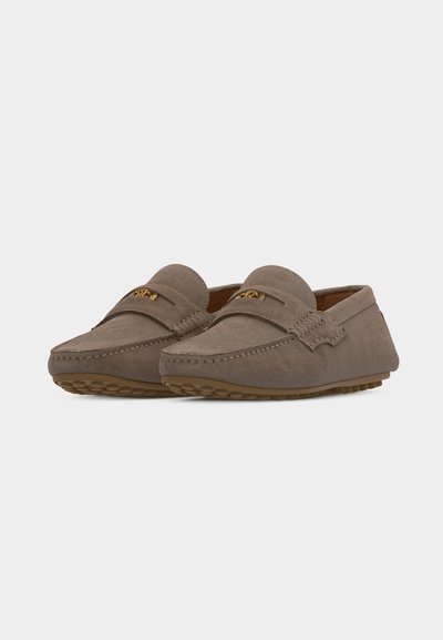 Paire de mocassins en daim taupe avec semelles en caoutchouc cloutées et détail en métal doré sur le dessus, présentée en léger angle sur fond blanc.