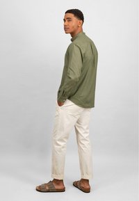 Camicia verde oliva a maniche lunghe, pantaloni chino chiari e sandali a infradito marroni. La camicia ha un colletto classico e dettagli di apertura sul retro.