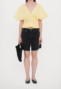 Top corto a pieghe giallo con scollo a V, abbinato a pantaloni shorts in denim neri, caratterizzati da orli sfrangiati. Borsa intrecciata nera e ballerine nere.