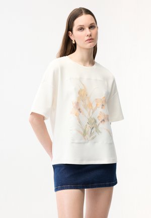 Nuori nainen, jolla on pitkät ruskeat hiukset, pukeutuneena valkoiseen ylisuurikokoiseen T-paitaan, jossa on pastellivärinen kukkakuosi, ja tummansiniseen farkkuhameeseen.