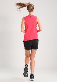 Röd ärmlös atletisk topp med rund halsringning och svart logotyp, i kombination med svarta shorts som har vita sidostrecker och atletiska skor.
