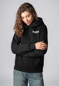 Schwarzer Kapuzenpullover mit einer vorderen Kängurutasche und einem bedruckten Logo auf der Brust. Hergestellt aus weichem Stoff mit gerippten Bündchen und Saum.