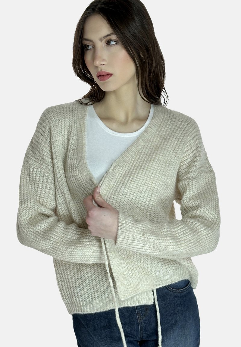 Beige gebreide cardigan met een V-hals, lange mouwen en een strikdetail aan de voorkant. Zachte textuur met een relaxte pasvorm. Gekocht over een witte top.