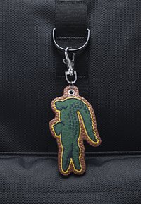 Leder-Schlüsselanhänger in Form eines grünen Alligators mit gelben Akzenten, verfügt über einen strukturierten braunen Rand, befestigt an einer schwarzen Tasche mit einem Metallclip.