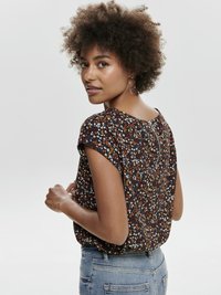 Käärus juustega naine, kellel on kõrvas rõngaskõrvarõngad, lühikeste varrukatega lilleline pluus ja sinised teksad, pööratud veidi seljaga ja vaatab üle õla.