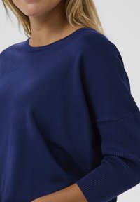 Pull bleu marine en tissu tricoté doux, doté d'un col rond et de poignets côtelés. Le design met l'accent sur une coupe confortable et une texture agréable.