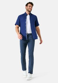 Korte mouwen blauwe linnen overhemd, witte T-shirt eronder, gecombineerd met marineblauwe broek en witte sneakers. Model loopt tegen een effen achtergrond.